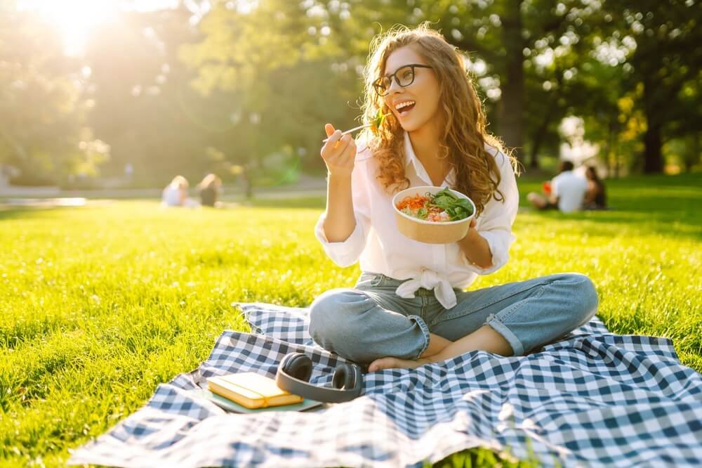 a waman is outside in the park and eating vagetable in a bawl